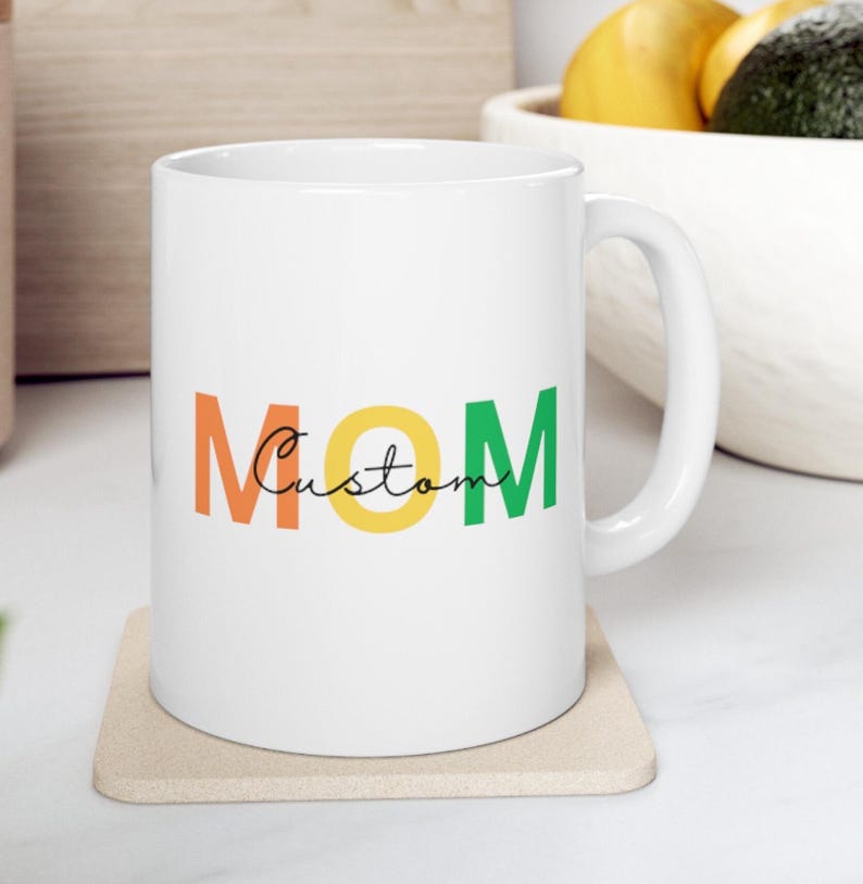 May include: White ceramic mug with the word "MOM" in orange, yellow, and green block letters. The word "Custom" is written in a cursive font between the letters. The mug sits on a beige coaster.