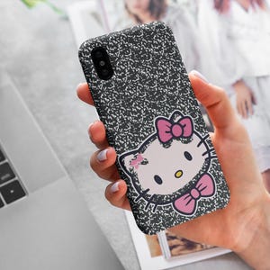 以下が含まれることがあります： 黒と白の電話ケース。ノート用紙のデザインとハローキティのグラフィック。ハローキティのグラフィックはピンクのリボンとピンクの蝶ネクタイが特徴です。電話ケースは人の手で持たれています。