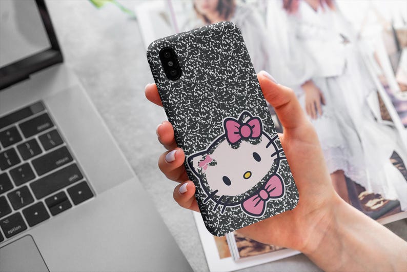 以下が含まれることがあります： 黒と白の電話ケース。ノート用紙のデザインとハローキティのグラフィック。ハローキティのグラフィックはピンクのリボンとピンクの蝶ネクタイが特徴です。電話ケースは人の手で持たれています。