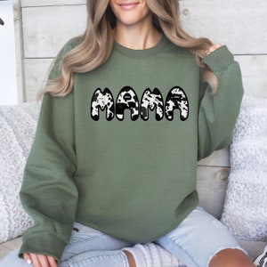 May include: An olive green sweatshirt with the word "MAMA" in large, bold letters across the front. The letters have a black outline and a cow print pattern in white and black. The sweatshirt has long sleeves and a crew neck.
