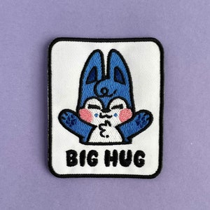 May include: Embroidered patch featuring a blue cartoon character with open arms, pink cheeks, and a kissy face. The patch is rectangular with rounded corners and the words "BIG HUG" in black text on a white background.