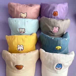 Pode incluir: Nove bon&eacute;s de beisebol de diferentes cores com rostos de animais bordados. Os bon&eacute;s s&atilde;o de v&aacute;rias cores, incluindo rosa, azul, amarelo, cinza e branco. Cada bon&eacute; tem um rosto de animal diferente bordado na frente, incluindo um gato, um urso, um coelho, um p&aacute;ssaro e um peixe.
