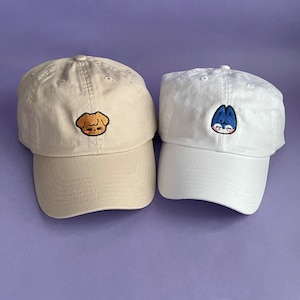 Left: Stone dad cap with Puppym (brown puppy) Skzoo embroidered on the center. Right: White dad hat with Wolf Chan (blue & white wolf) Skzoo embroidered on the center.