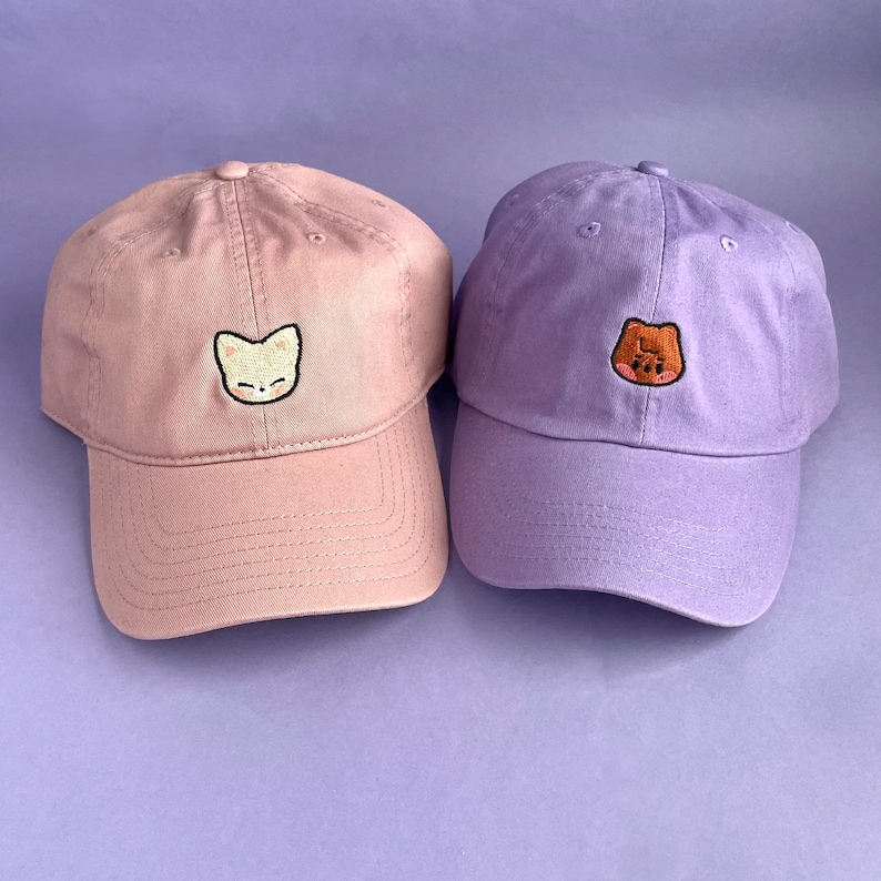 Left: Pink dad cap with FoxINy (beige fox) Skzoo embroidered on the center. Right: Lavender dad hat with Han Quokka (brown quokka) Skzoo embroidered on the center.