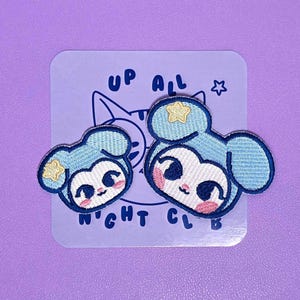 Puede incluir: Dos parches bordados con un personaje de dibujos animados con orejas azul claro, cara blanca, ojos oscuros y mejillas rosadas. Cada parche tiene una pequeña estrella amarilla en una oreja. Se muestran sobre una tarjeta con el texto UP ALL NIGHT CLUB.