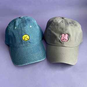Left: Navy dad cap with Bbokari (yellow chick) Skzoo embroidered on the center. Right: Gray dad hat with Dwaekki (pink pig bunny) Skzoo embroidered on the center.
