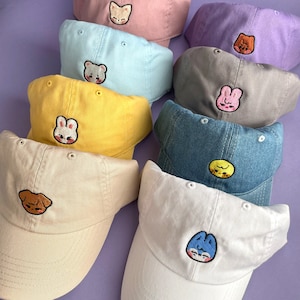 Pode incluir: Nove bon&eacute;s de beisebol bordados em v&aacute;rias cores com diferentes desenhos de animais. Os bon&eacute;s est&atilde;o dispostos em semic&iacute;rculo sobre um fundo roxo.