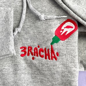 May include: Grey hoodie with a drawstring and a front pocket. The hoodie features an embroidered red and green sriracha bottle design and the word "3RACHA" in red embroidery.