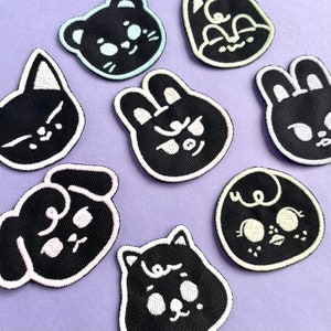 May include: Eight black embroidered iron-on patches featuring various animal faces with white outlines. The animals include a cat, a dog, a bunny, and a bear.