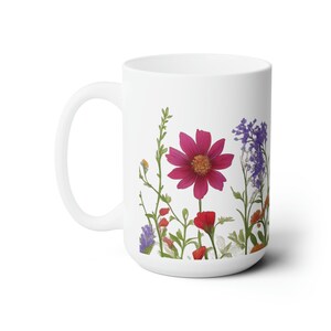 May include: White ceramic mug featuring a vibrant floral design. The design includes a large magenta flower, red poppies, and purple wildflowers, with green stems and leaves. The mug has a curved handle.