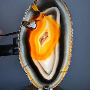 Op de afbeelding: Een verlichte, ovaalvormige agaatplak met concentrische banden van doorschijnend oranje, amber en diepzwarte kleuren. Het midden toont een heldere, gloeiende kern met een kleine kristallijne insluiting, ondersteund door een metalen standaard.