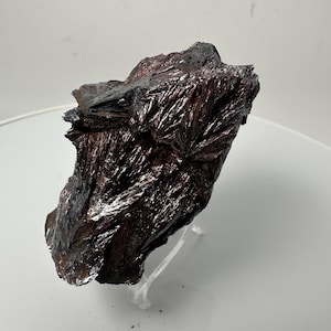 May include: A large, dark gray, iridescent, mineral specimen with a rough, textured surface. The mineral is on a clear plastic stand.