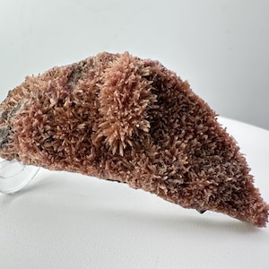 May include: A natural mineral specimen featuring a cluster of small, reddish-brown crystals. The crystals have a spiky, textured appearance, and the mineral sits on a clear display stand. The overall shape is triangular.