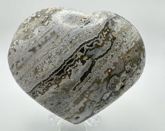 May include: A heart-shaped polished stone with a grey, white and brown banded pattern. The stone has a smooth, polished surface.