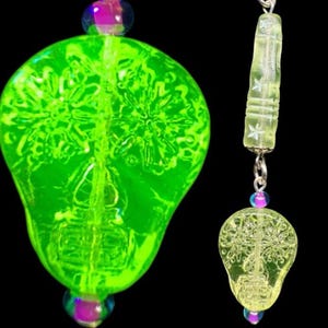Puede incluir: Un colgante de calavera de vidrio verde neón vibrante con intrincados grabados florales en la superficie. La calavera cuelga de una disposición vertical de una cuenta cilíndrica con grabados de estrellas y una pequeña cuenta de vidrio iridiscente.