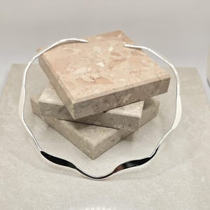 May include: A silver-colored, wavy collar necklace. The necklace is displayed on a stack of three square, textured, beige and pink stone tiles. The necklace has a smooth, reflective surface and a unique, organic shape.