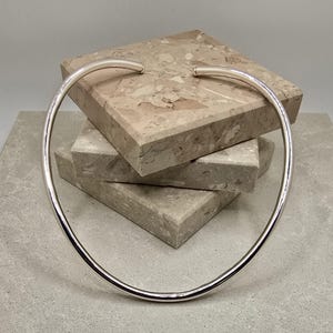 May include: A polished silver choker necklace with a minimalist design. The necklace is a smooth, curved band that sits around the neck. The ends of the necklace are slightly curved. The necklace is displayed on a stack of stone blocks.