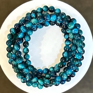 May include: A collection of teal and blue beaded bracelets arranged in a circular pattern. The beads have a polished, reflective surface, and the color transitions from dark teal to bright blue. The bracelets are displayed on a white surface.