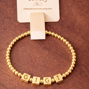 May include: A gold-toned beaded bracelet featuring small round metallic beads and four square letter beads spelling GIGI. The bracelet is attached to a beige display card printed with The Bead Hive Society and the website address.