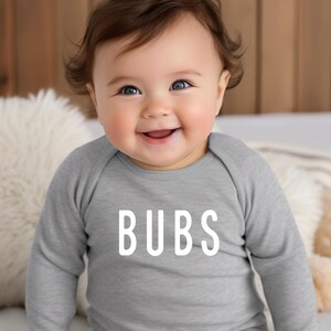 May include: A smiling baby wearing a gray long-sleeved bodysuit with the word "BUBS" in white block letters. The baby has brown hair and blue eyes.