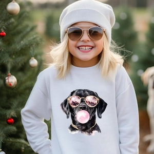 May include: A white sweatshirt featuring a graphic of a black dog wearing sunglasses and blowing a bubblegum bubble. The dog has a gold chain collar. The person is wearing a white beanie and sunglasses.