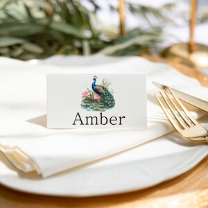 May include: A white place card with a peacock illustration and the name "Amber" printed in black.