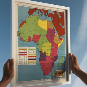 May include: A framed vintage map of Africa, featuring a colorful political division with countries labeled. The map is set against a light blue background and held by two hands. The frame is white.