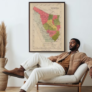 Op de afbeelding: Een vintage kaart van de staat Sonora, Mexico, ingelijst in een donkerhouten lijst, hangt aan een witte muur. Een persoon zit op een witte bank met een houten frame, gekleed in een bruin shirt en een witte broek.