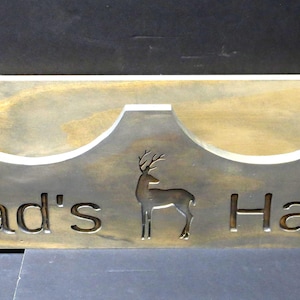 May include: A wooden hat rack with two compartments, each shaped like a curved half-circle. The rack is painted in a light brown color with a distressed finish. The words "Dad's Hats" are engraved on the front of the rack, along with a silhouette of a deer.