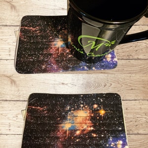 May include: Two black and white galaxy print coasters with a black mug on top of one of the coasters. The mug has a green logo that says "M's Crafts".