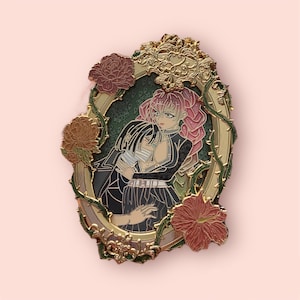 Puede incluir: Pin de esmalte dorado con una pareja abrazándose dentro de un marco decorativo. El marco está adornado con detalles florales y enredaderas. La pareja se representa en una pose romántica, con la mujer vistiendo un vestido rosa y el hombre un traje negro.