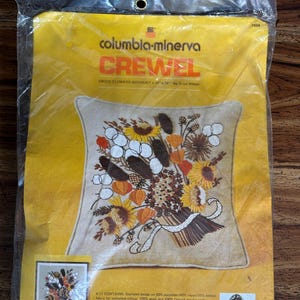 May include: A Columbia-Minerva crewel embroidery kit for a 16 inch by 16 inch pillow featuring a dried flower bouquet design by Erica Wilson. The yellow packaging displays the finished project, materials list, and a portrait of the designer.