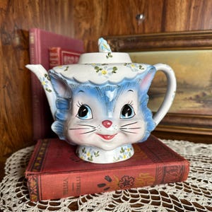 May include: A ceramic teapot shaped like a cat with blue fur, large black eyes, a pink nose, and a red mouth. The teapot features a white lid with a small blue bird handle and is decorated with painted blue and yellow floral patterns.