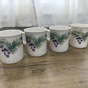 May include: A set of four off-white ceramic mugs, each featuring a painted design of purple grape clusters and green vine leaves encircling the exterior. The mugs have a smooth, rounded handle and a simple, cylindrical shape with a flared rim.