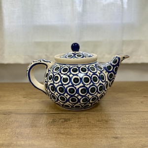 May include: A ceramic teapot featuring a cream-colored base with a repeating pattern of dark blue and teal circular motifs. The teapot has a rounded body, a curved handle, a tapered spout, and a matching lid with a small, solid blue knob.