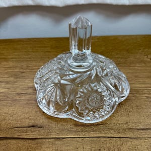 May include: A clear glass lid featuring a faceted, pointed handle and a decorative pressed pattern of starbursts and geometric shapes. The circular lid has a scalloped edge and a reflective, transparent surface with intricate cut glass detailing.