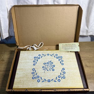 May include: A rectangular electric warming tray with a gold-toned surface featuring a blue floral pattern in a central wreath design. The tray includes a white power cord and a small rectangular guarantee card from Atlantic Precision Works Inc.