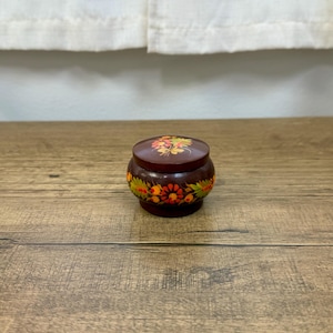May include: A small, round, dark brown wooden lidded box featuring hand-painted floral motifs in shades of red, orange, yellow, and green. The lid and the base of the container are decorated with detailed botanical patterns and foliage.