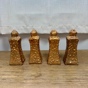 May include: Four identical salt and pepper shakers with a metallic gold finish and a textured, irregular surface. Each shaker features a tapered, rectangular body with a rounded top and a small hole for dispensing, standing upright in a row.
