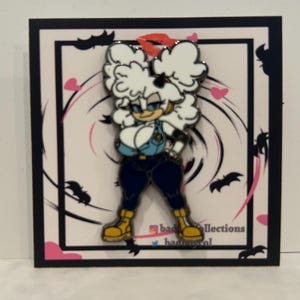 May include: Enamel pin featuring a cartoon character with white hair, blue eyes, and a blue outfit. The pin is set against a black and white background with bats and hearts. The pin is on a black square.