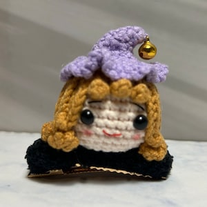 May include: A small crocheted doll with blonde hair, wearing a purple hat with a gold bell, and a black dress. The doll is smiling and has big eyes.