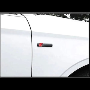 May include: A black and red S line emblem attached to a white car. The emblem is rectangular with the letters "S line" in a thin, sans-serif font. The "S" is red, and the rest of the text is black.
