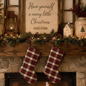 Puede incluir: Dos calcetines navideños con un patrón de cuadros rojos, blancos y negros cuelgan de una repisa de madera. Sobre la repisa, un letrero dice Have yourself a merry little Christmas, flanqueado por una linterna, un árbol, una casa y una jarra.