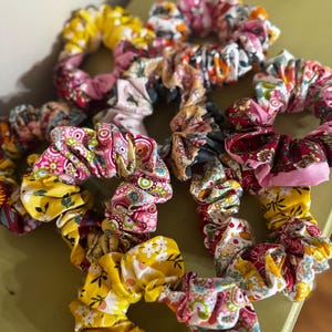 May include: A collection of fabric hair scrunchies featuring various colorful patterns including yellow floral prints, pink paisley designs, and multi-colored botanical motifs. The scrunchies are gathered with elastic, creating a ruffled texture.