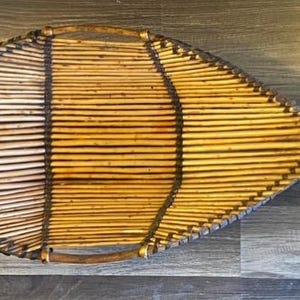 Può includere: Un vassoio da portata in vimini intrecciato a forma di pesce, in tonalità marroni e beige. Il vassoio è realizzato con materiali naturali e ha un aspetto rustico e artigianale.