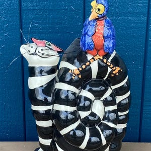 Könnte beinhalten: Eine schwarze und weiße gestreifte Keramik-Katzenfigur mit einem blauen und roten Vogel, der auf ihrem Rücken sitzt. Der Vogel hat eine goldene Krone und schaut nach rechts. Die Katze schaut nach oben und hat eine rosa Nase.