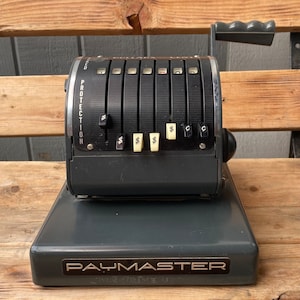 May include: A vintage gray metal cash register with a black lever and a silver label that reads "PAYMASTER".