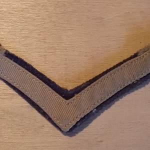 May include: A gray chevron patch with a dark blue border. The patch is made of fabric and has a woven texture.
