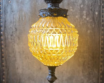 May include: A vintage, amber glass pendant light fitting with a black metal frame and a diamond-shaped pattern. The light is on and illuminating the room.