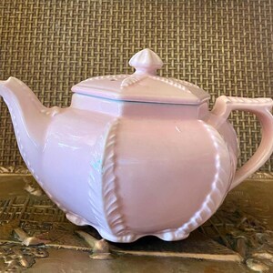 Puede incluir: Una tetera de cerámica rosa claro con tapa. La tetera tiene un pico y asa curvos, y un diseño en relieve decorativo alrededor del cuerpo. La tapa está rematada con un pequeño adorno. La tetera está sobre una bandeja decorativa.